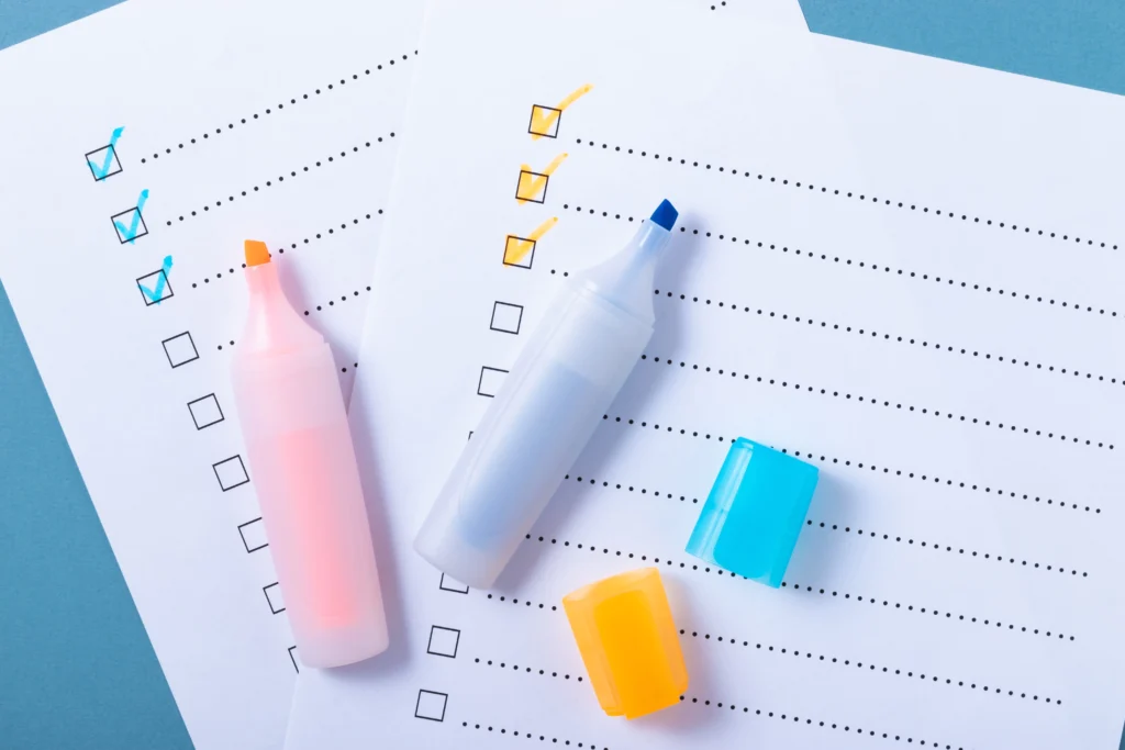 Two checklists with black outlined boxes and dotted lines lie on a blue surface. Some boxes are checked with blue and yellow highlighter marks. Two uncapped highlighters, one pink with an orange tip and one white with a blue tip, rest on the papers alongside their matching caps."